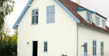 Hvid husfacade med blå vinduesrammer, rødt tegltag og matchende blåt havemøbel.