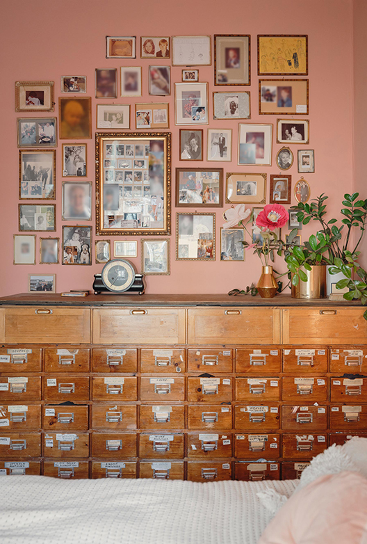 Rosa væg dækket af en samling indrammede fotografier og billeder over et stort vintage-skab med mange små skuffer.