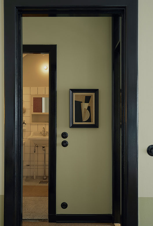 Hallway with light green walls, black door frames, and a small geometric artwork, with a view into the bathroom.