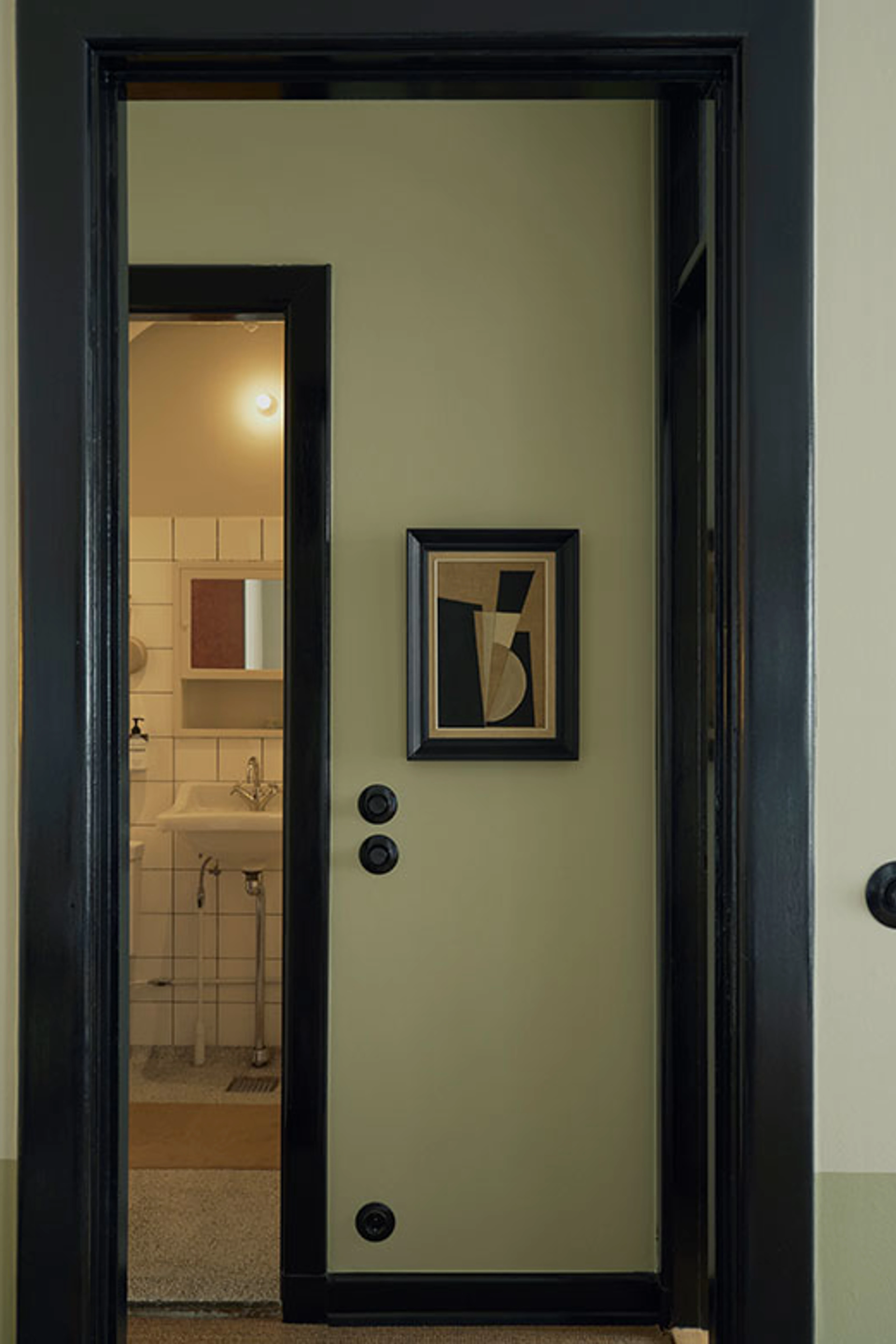 Hallway with light green walls, black door frames, and a small geometric artwork, with a view into the bathroom.