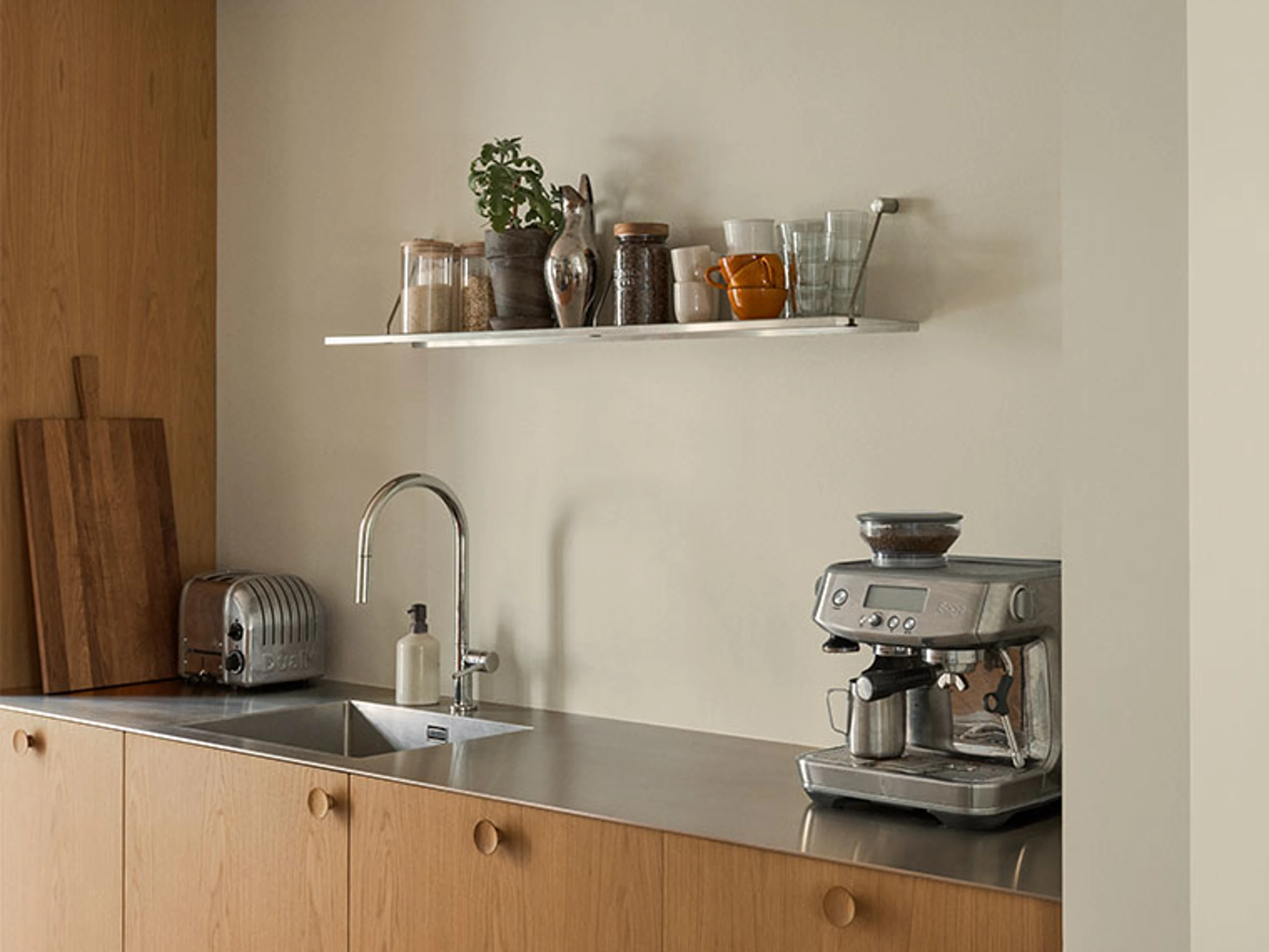 Køkkenbord med vask, kaffemaskine og åben hylde med planter, glas og kopper.