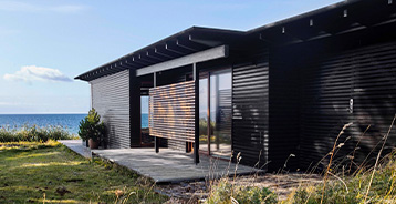 Moderne sommerhus med sortmalet træfacade og store glaspartier placeret tæt ved kysten. Huset er omgivet af marehalm og natur med havet i baggrunden.