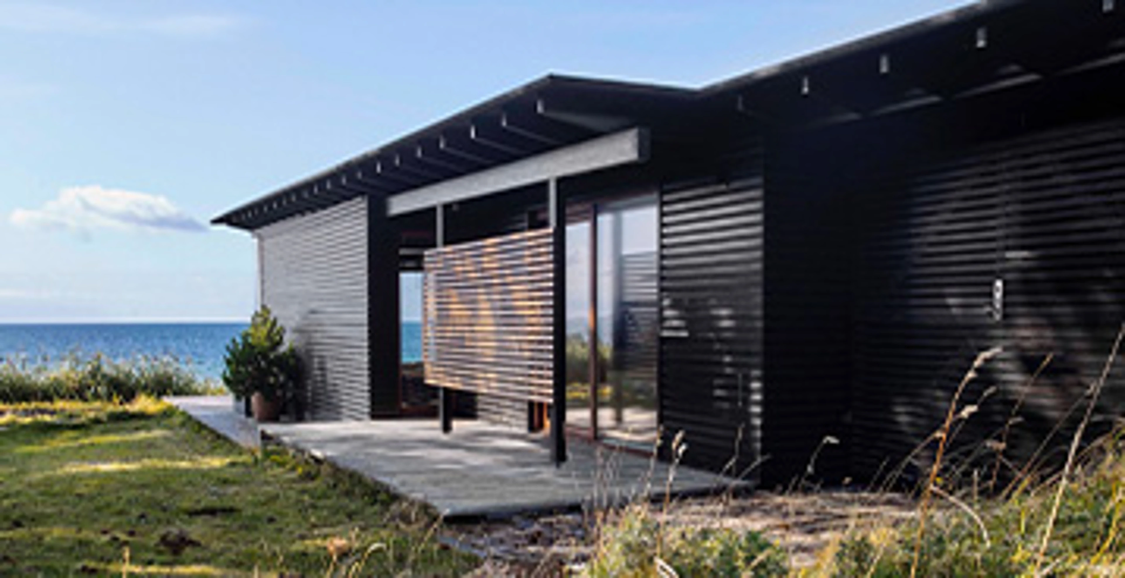 Moderne sommerhus med sortmalet træfacade og store glaspartier placeret tæt ved kysten. Huset er omgivet af marehalm og natur med havet i baggrunden.