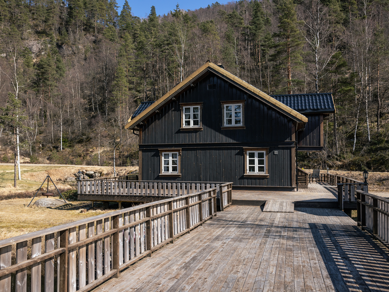 Svart trähus med en stor träaltan och skog i trädgården.