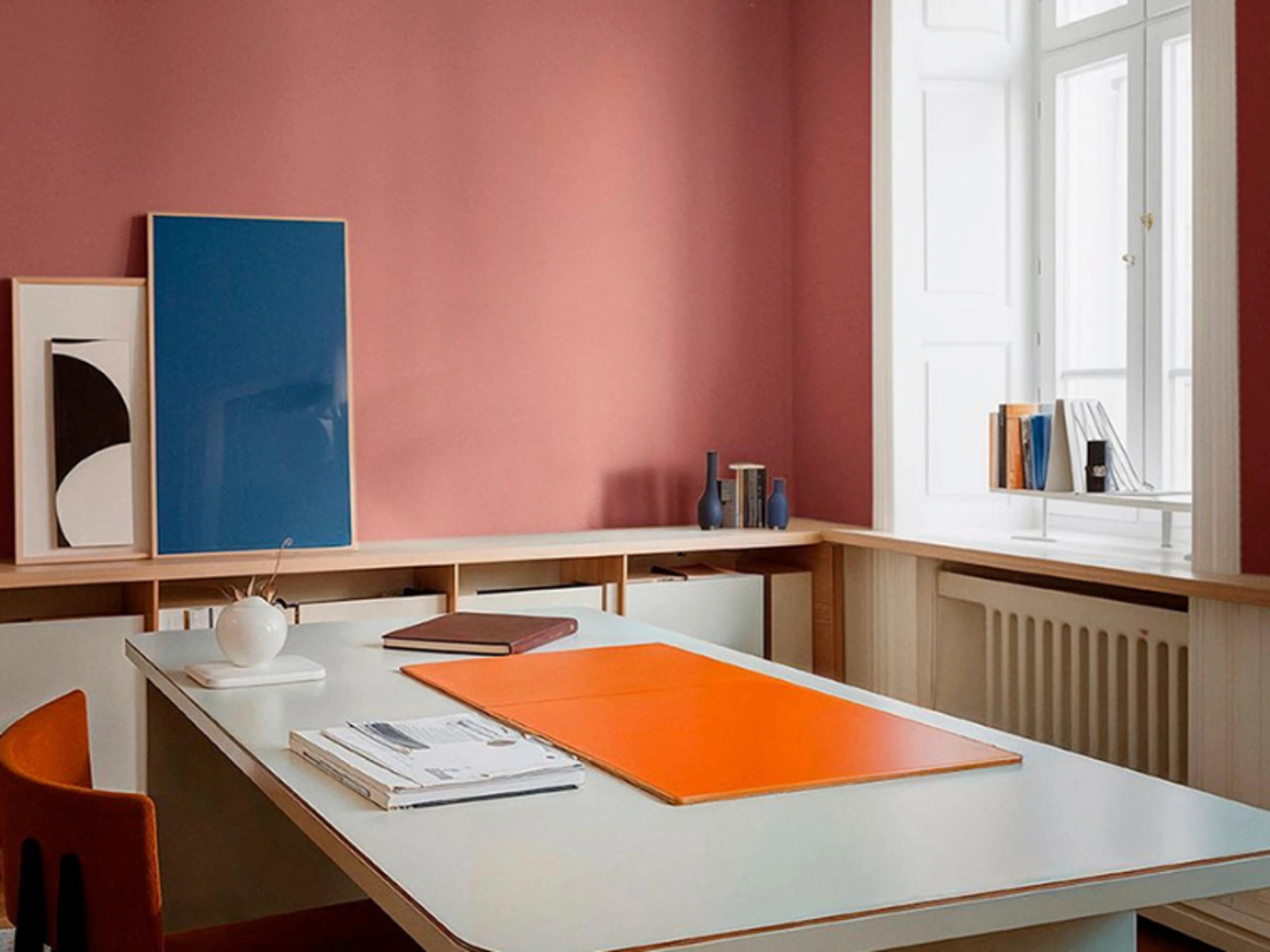 Kontor med rosamalt vegg, hvitt skrivebord med oransje skriveunderlag, et blått innrammet bilde og en oransje stol foran et høyt vindu.