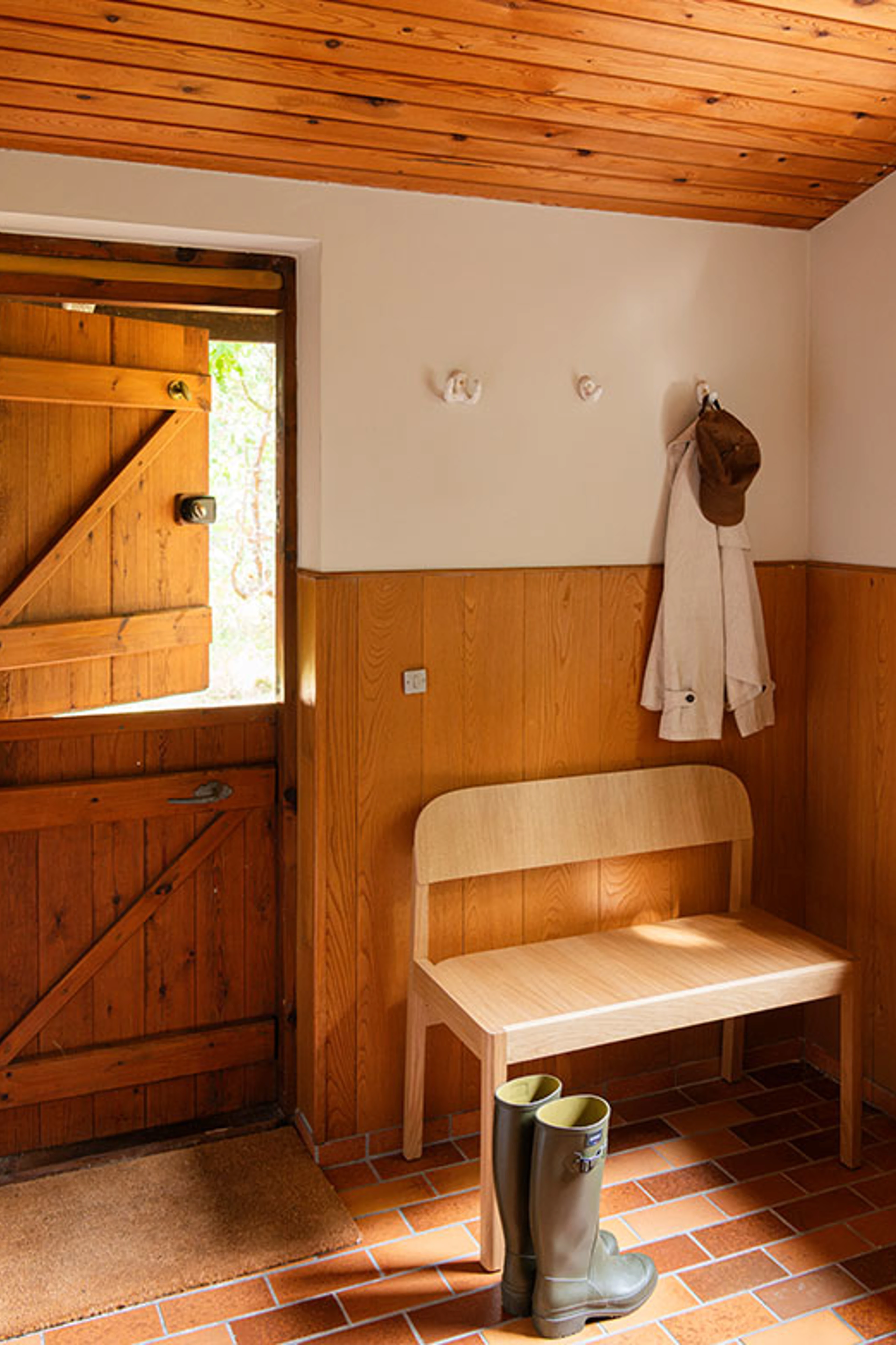 Hallway with wood panelling, a white wall and a wooden ceiling, with a light bench and rubber boots on terracotta tiles.