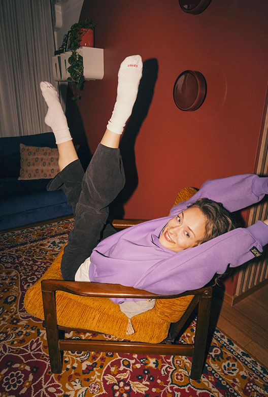 Kvinde i lilla sweatshirt ligger afslappet i en sennepsgul lænestol med benene løftet op ad den røde væg.