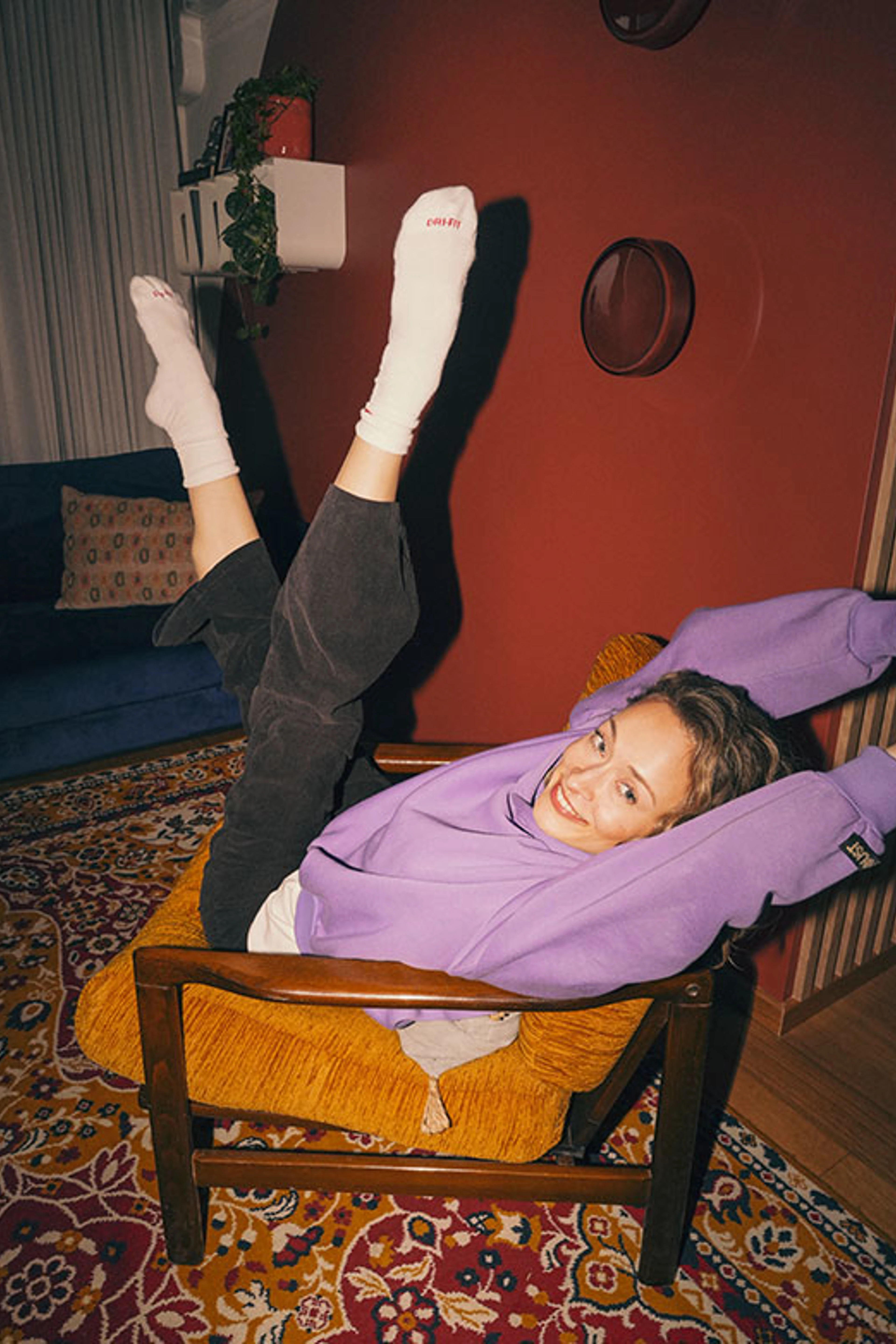 Kvinde i lilla sweatshirt ligger afslappet i en sennepsgul lænestol med benene løftet op ad den røde væg.