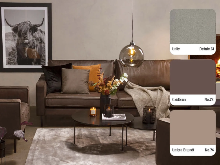 Living room with warm grey walls, a brown leather sofa and armchair, round black coffee tables, a glass pendant lamp and a framed Highland cattle photograph, with three colour swatches on the right: Unity Detale 61, Oxidbrun No.73 and Umbra Brændt No.74.