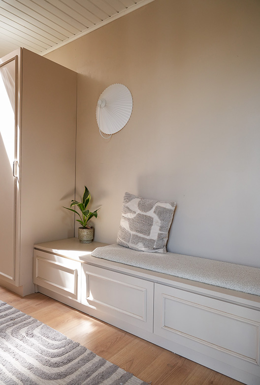 Entré med indbygget opbevaringsbænk, væg og skabe i varme beige nuancer. En grøn plante, en dekorativ pude og dagslys fra siden skaber et roligt og indbydende udtryk.