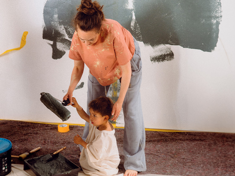 Person and child painting a wall together with a paint roller and paint tray during a home decorating project.