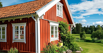 Rødmalet træhus med hvide vinduesrammer og gavldetaljer. Facaden er omgivet af grøn have og beplantning, som fremhæver husets klassiske nordiske udtryk og malede træværk.