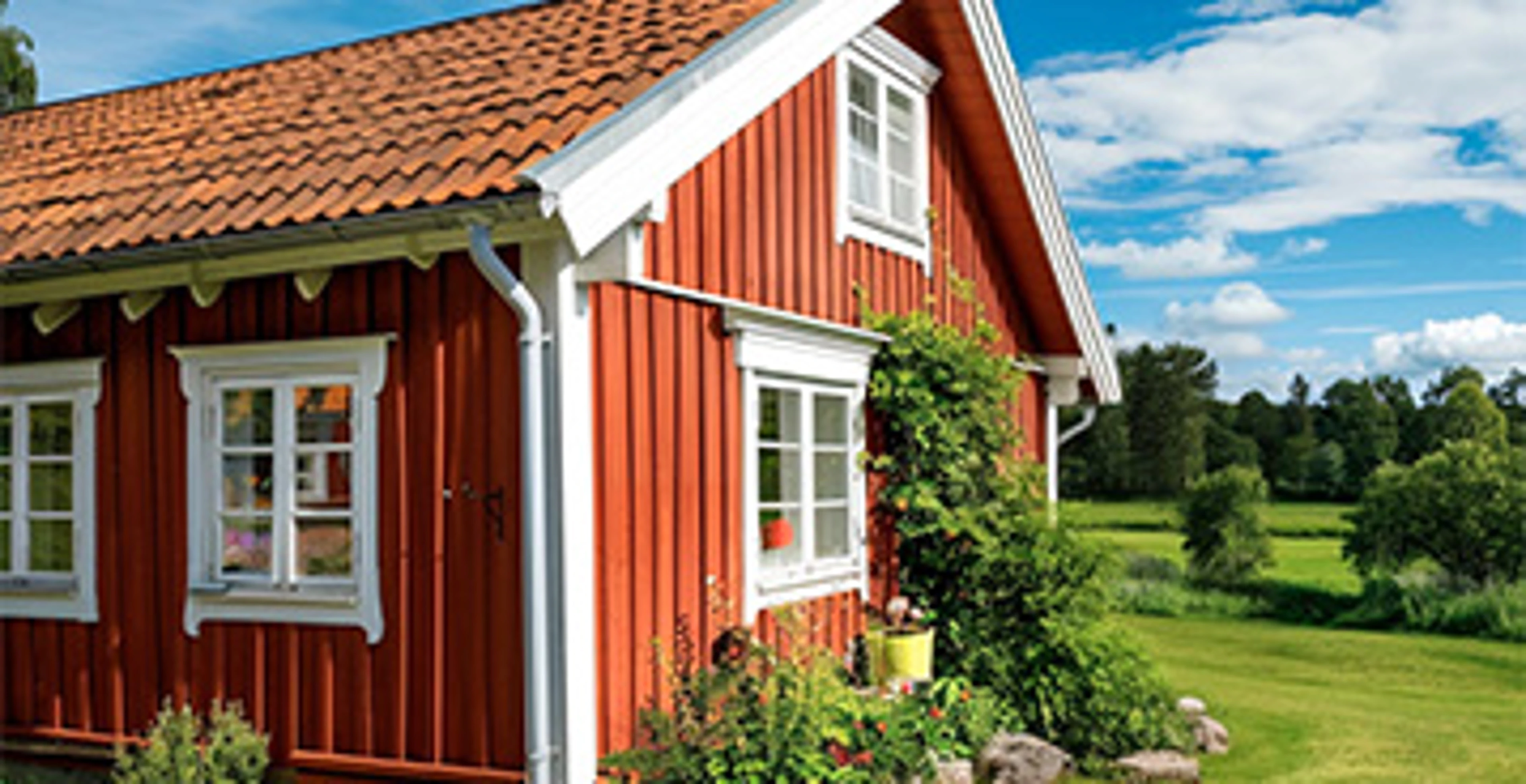 Rødmalet træhus med hvide vinduesrammer og gavldetaljer. Facaden er omgivet af grøn have og beplantning, som fremhæver husets klassiske nordiske udtryk og malede træværk.