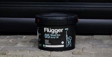 Et spann Flügger WoodTex 05-tremaling står på brostein foran en svartmalt trevegg.