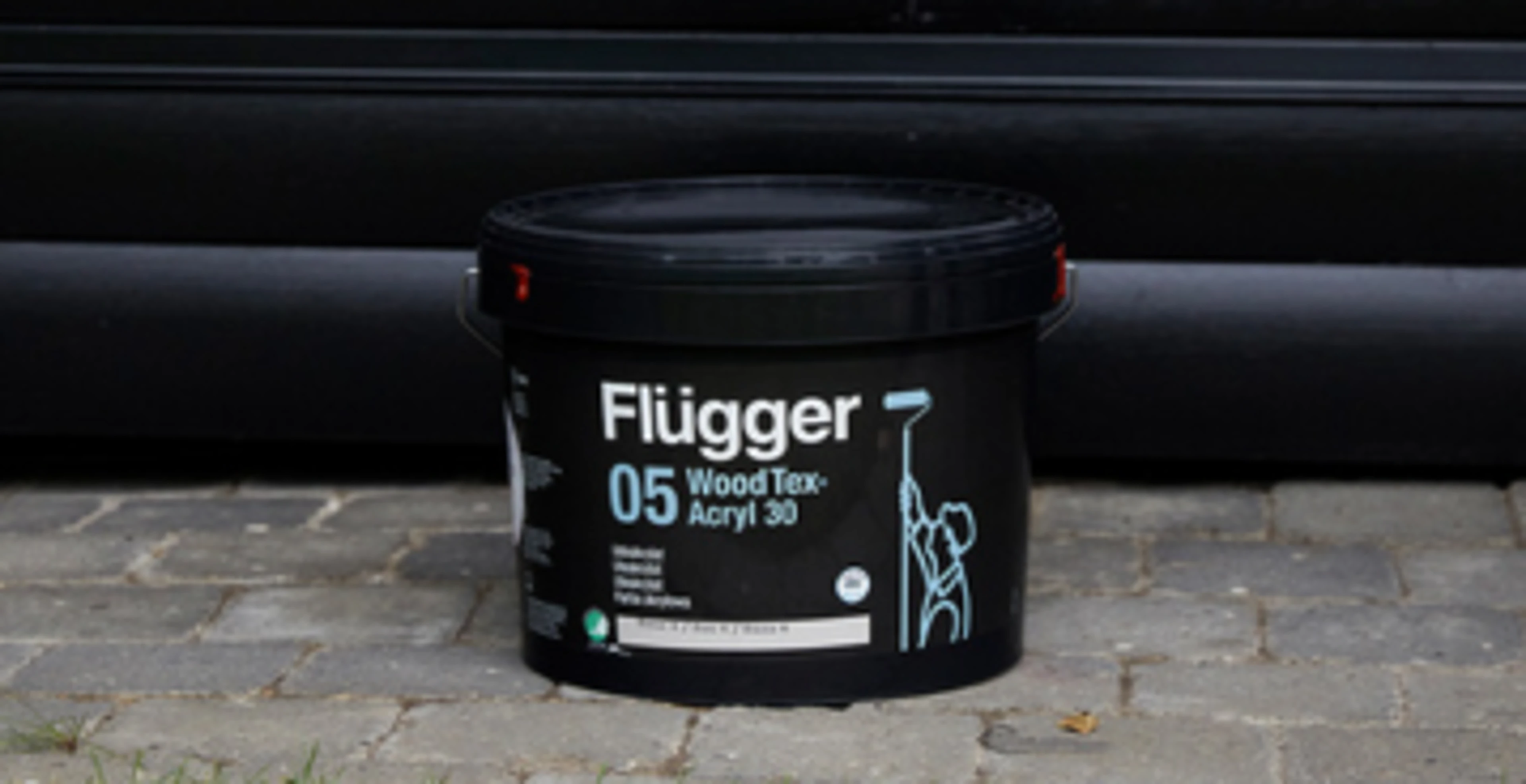 Et spann Flügger WoodTex 05-tremaling står på brostein foran en svartmalt trevegg.