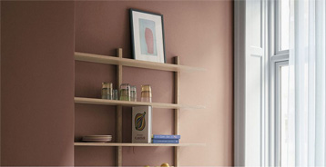 Corner of a bright room with walls painted in the warm terracotta colour Sienna FL8064. A light wooden shelf with books and ceramics, a large window with white curtains and light panels create an inviting and harmonious atmosphere.