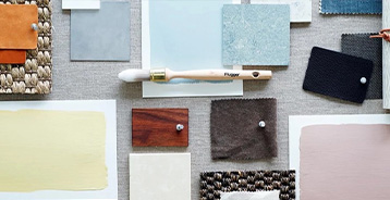 Materialeboard med farveprøver, tekstiler, træprøver og en pensel arrangeret på en lys overflade. Farvefelter i bløde grønne, beige og rosa nuancer indgår i sammensætningen.