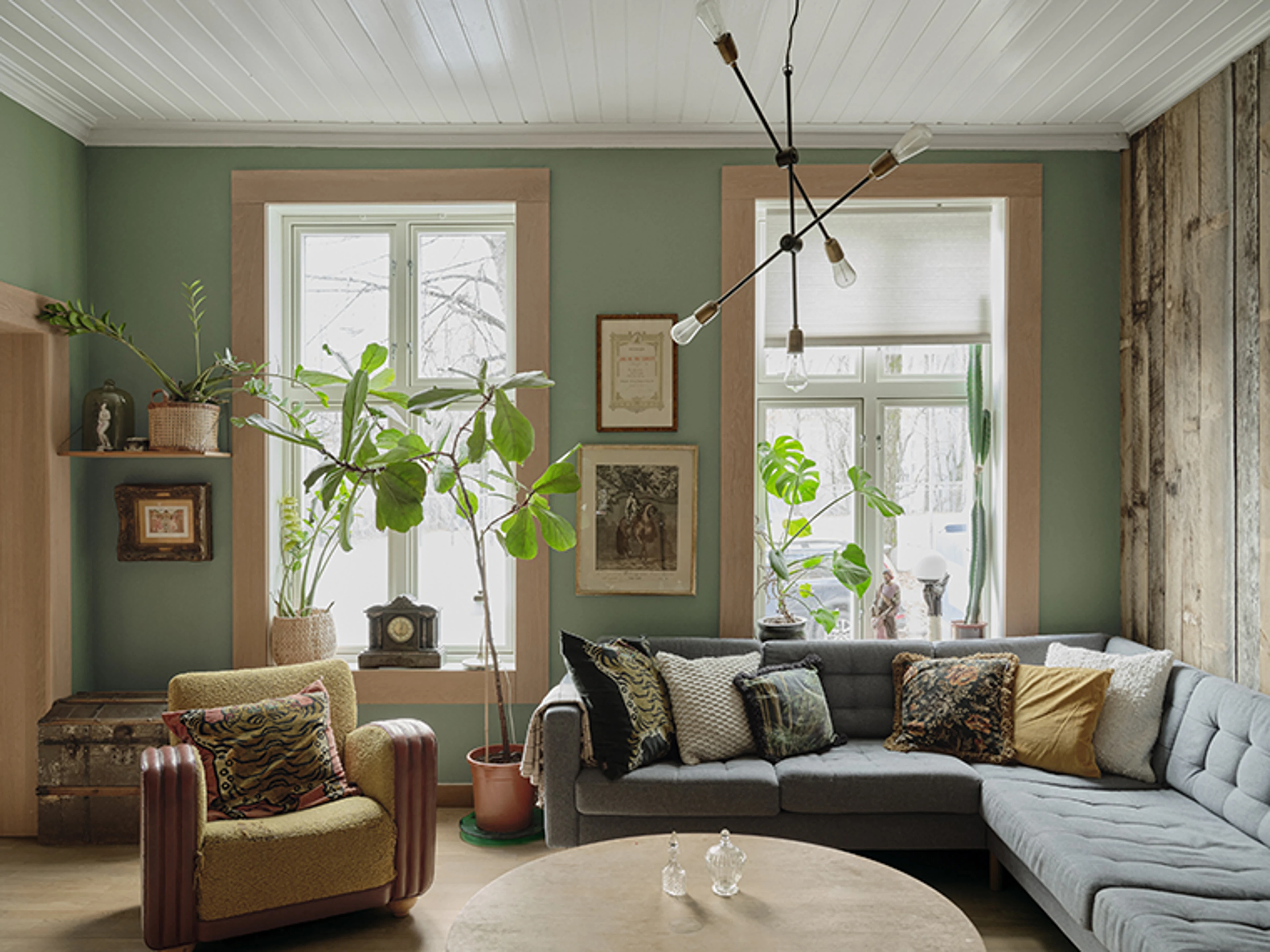 Grøn stue med store vinduer, mange planter, grå hjørnesofa og en sennepsgul vintage-lænestol.