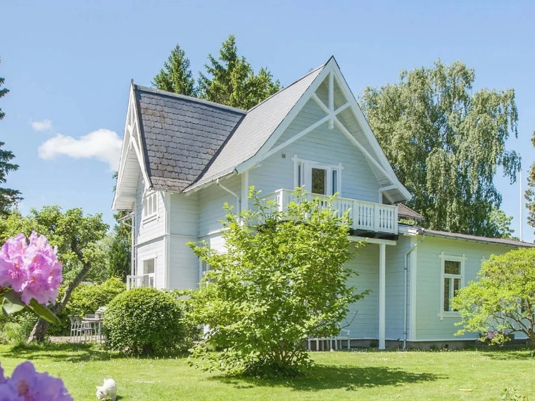 Ljusblå trävilla med vita lister, en balkong och ett skifferttak, omgiven av en grön trädgård och höga träd.