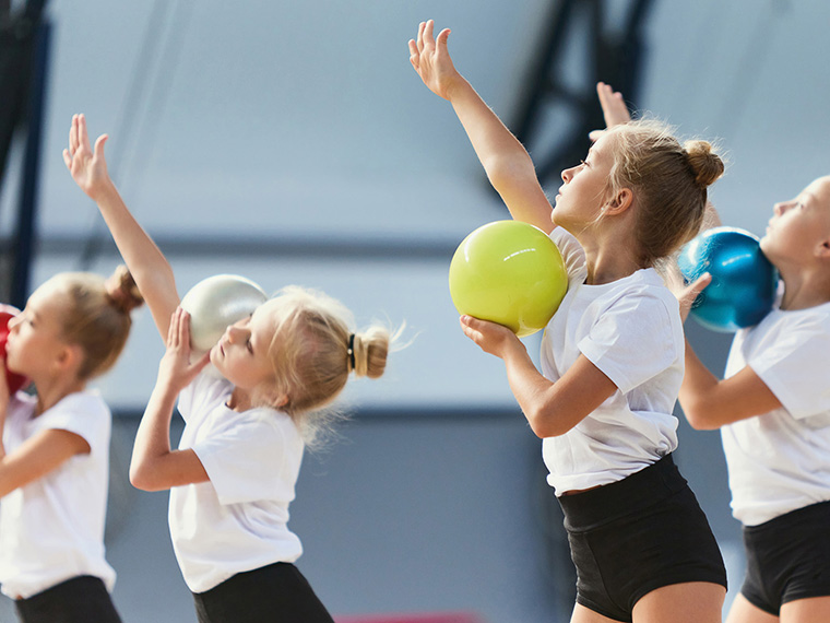 Flere børn træner rytmisk gymnastik i en idrætshal. Børnene holder farvede gymnastikbolde og udfører en koordineret øvelse med armene løftet. Hallens lyse baggrund fremhæver bevægelse og aktivitet.