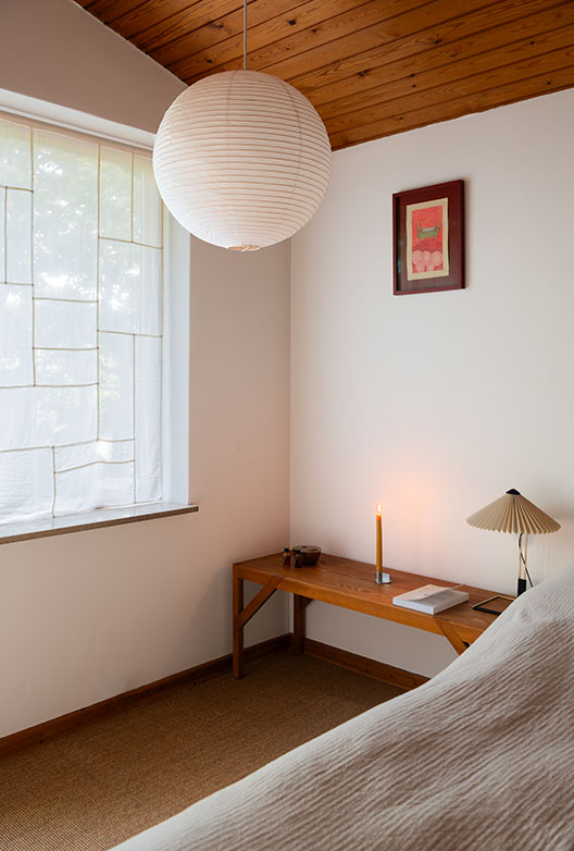 Bedroom with white walls, a wooden ceiling and a large rice paper lamp above a wooden bench with a lit candle.