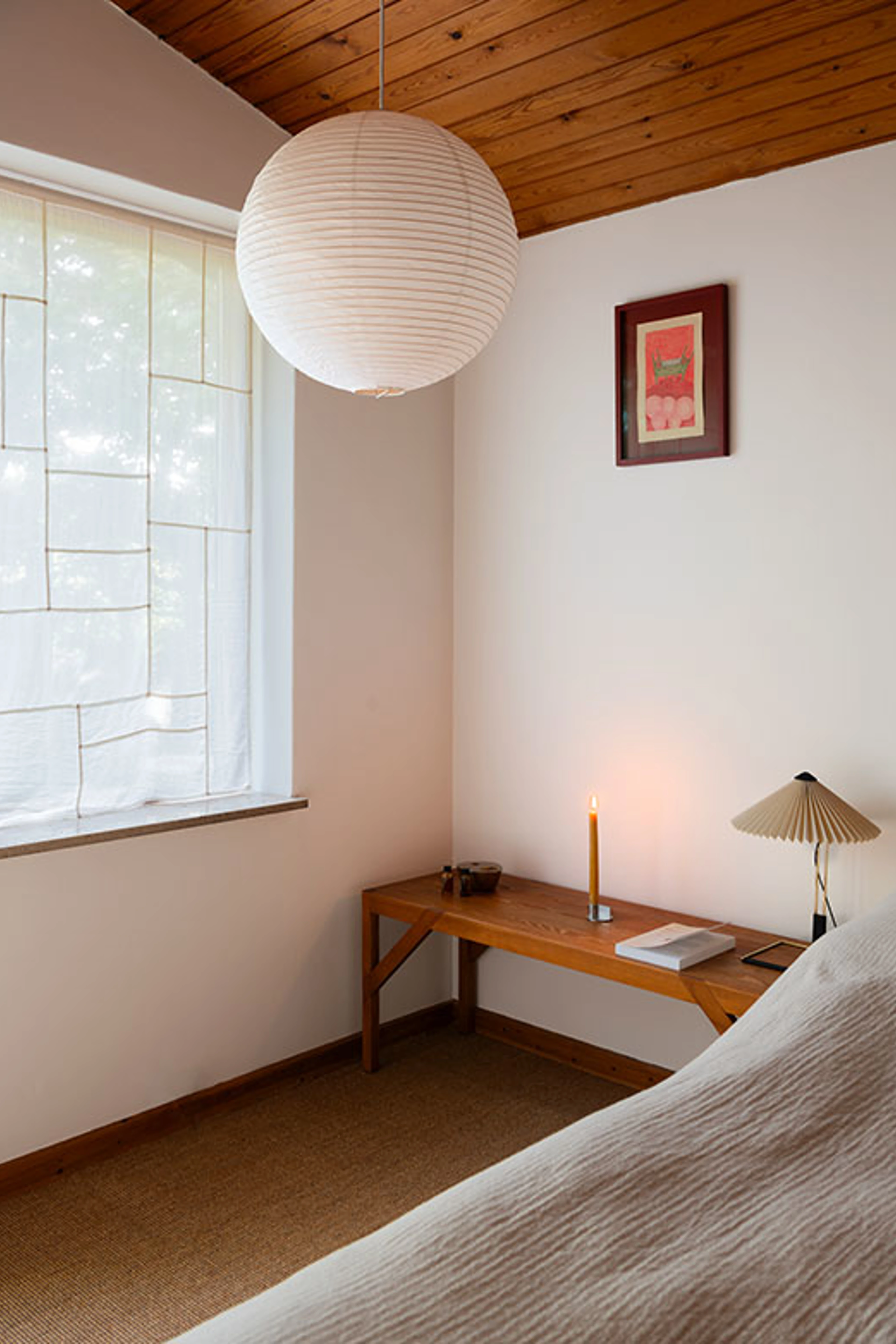 Bedroom with white walls, a wooden ceiling and a large rice paper lamp above a wooden bench with a lit candle.