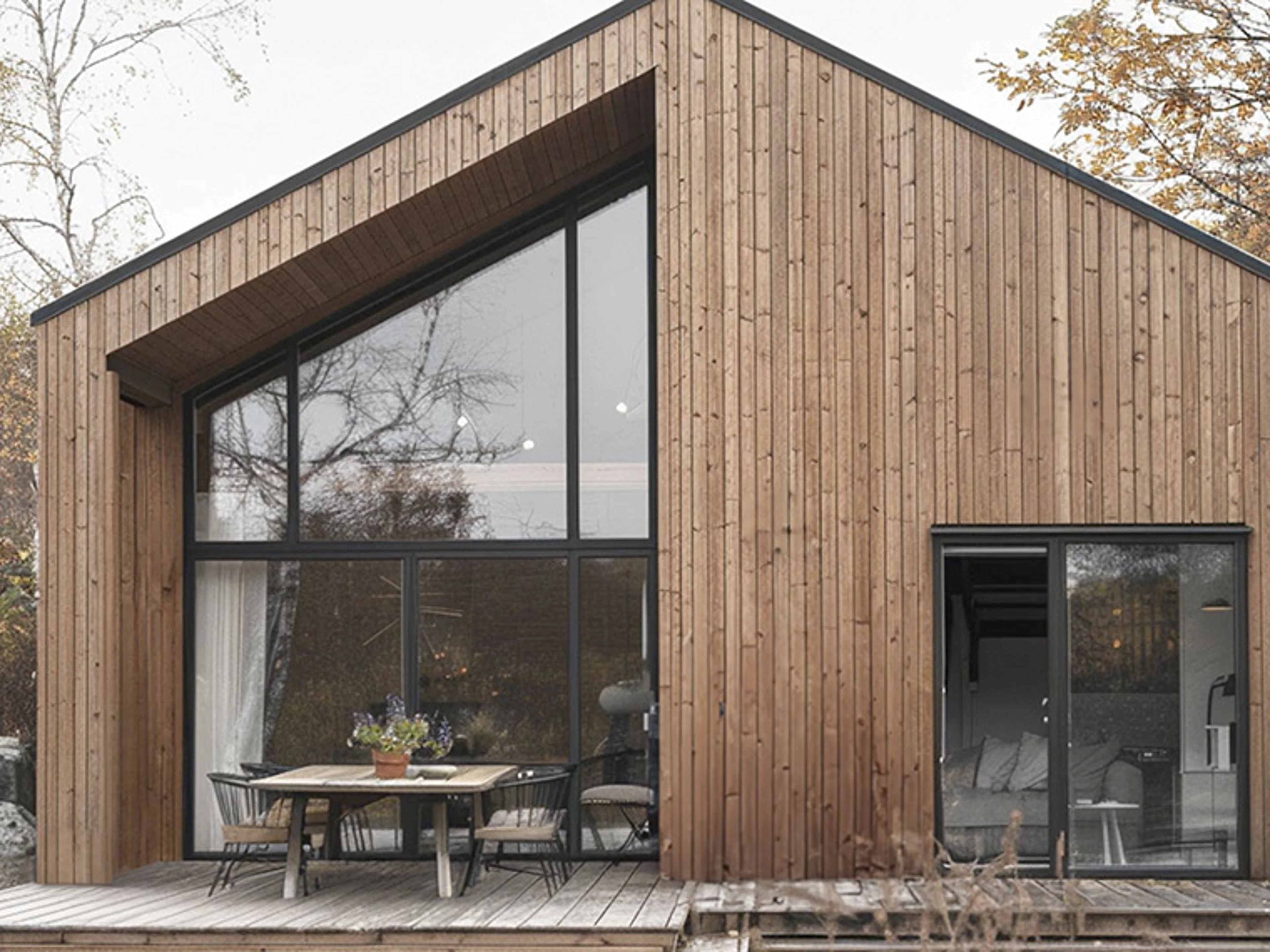 Moderne hus med facade i naturligt træ og store gulv-til-loft-vinduer. Foran huset ligger en træterrasse med spisebord og stole.