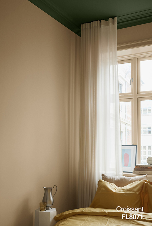 Sovrum med väggar målade i den varma beige färgen Croissant FL8071. Taket är målat mörkgrönt, och ljusa gardiner, sängkläder i okergula nyanser och dagsljus från ett stort fönster skapar en lugn och mysig atmosfär.
