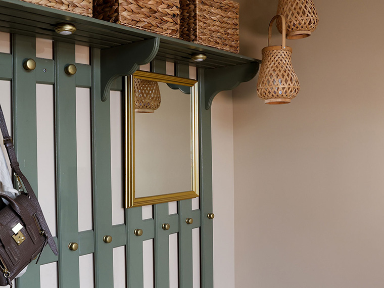 Garderobe med beige vægge, og en grøn knagerække med et spejl i guldramme. På hattehylden er der kurve i flettet materiale.