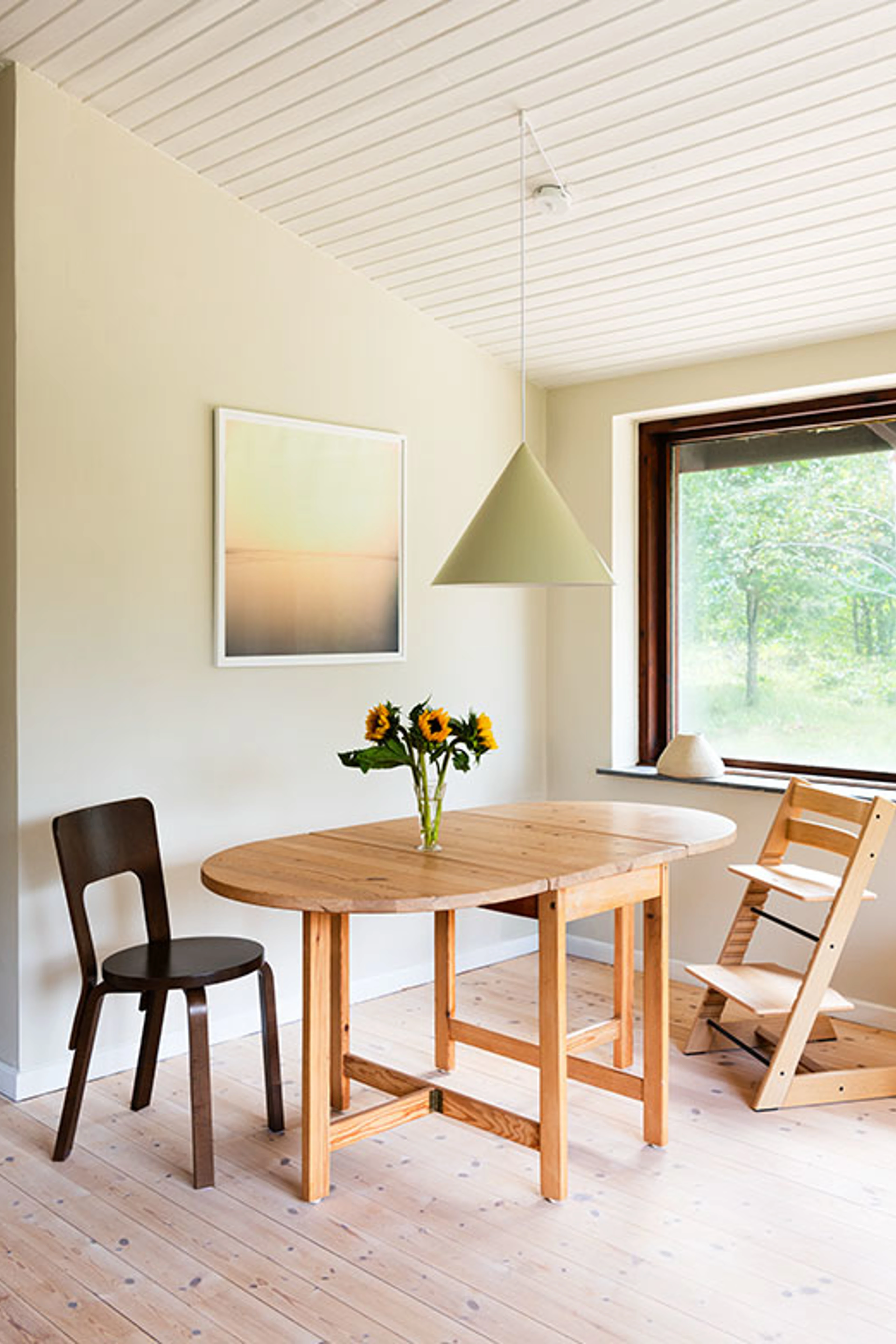 Dining area with light beige walls, a pine dining table with sunflowers and a green pendant lamp.