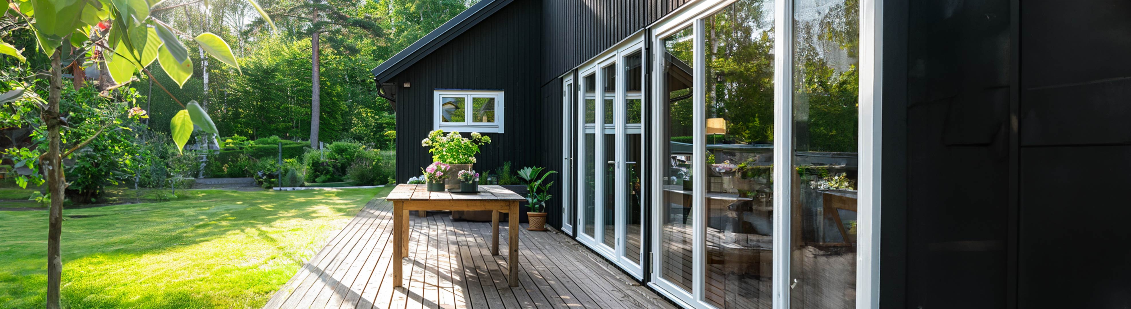 Sortmalet træhus med stor træterrasse og glaspartier ud mod en grøn have. På terrassen står et træbord med blomster.