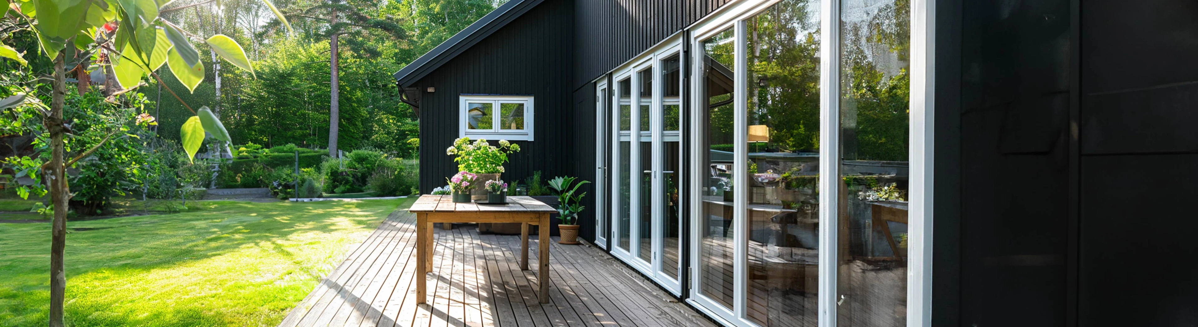 Sortmalet træhus med stor træterrasse og glaspartier ud mod en grøn have. På terrassen står et træbord med blomster.