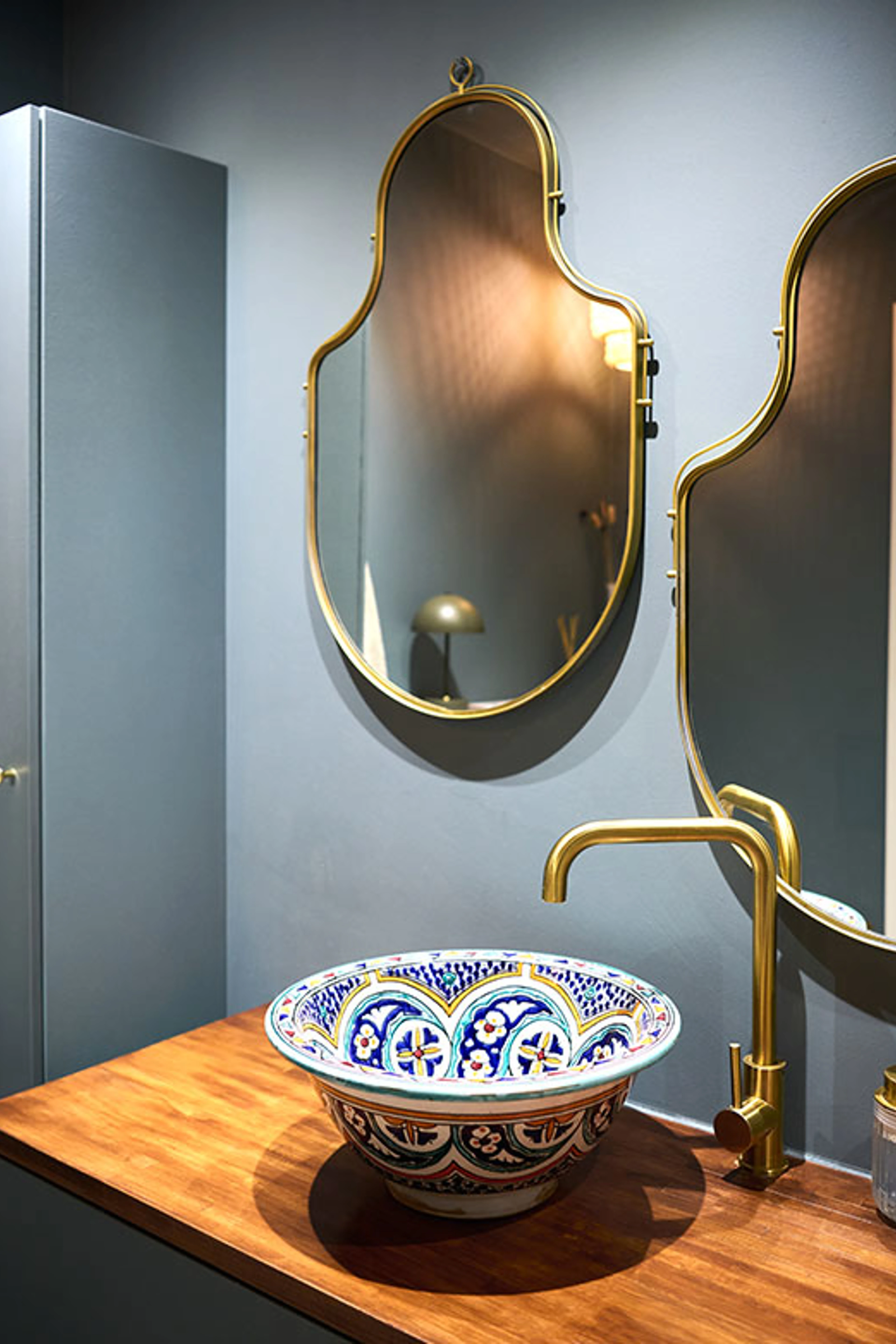 Bathroom with a dark blue-gray wall, decorative patterned sink, wooden countertop, brass faucet, and arched mirrors.