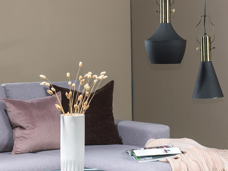 Et hjørne av en stue med en lys sofa, puter i rosa og mørke nyanser, en hvit vase med tørkede grener samt to mørkeblå pendellamper foran en beige vegg.