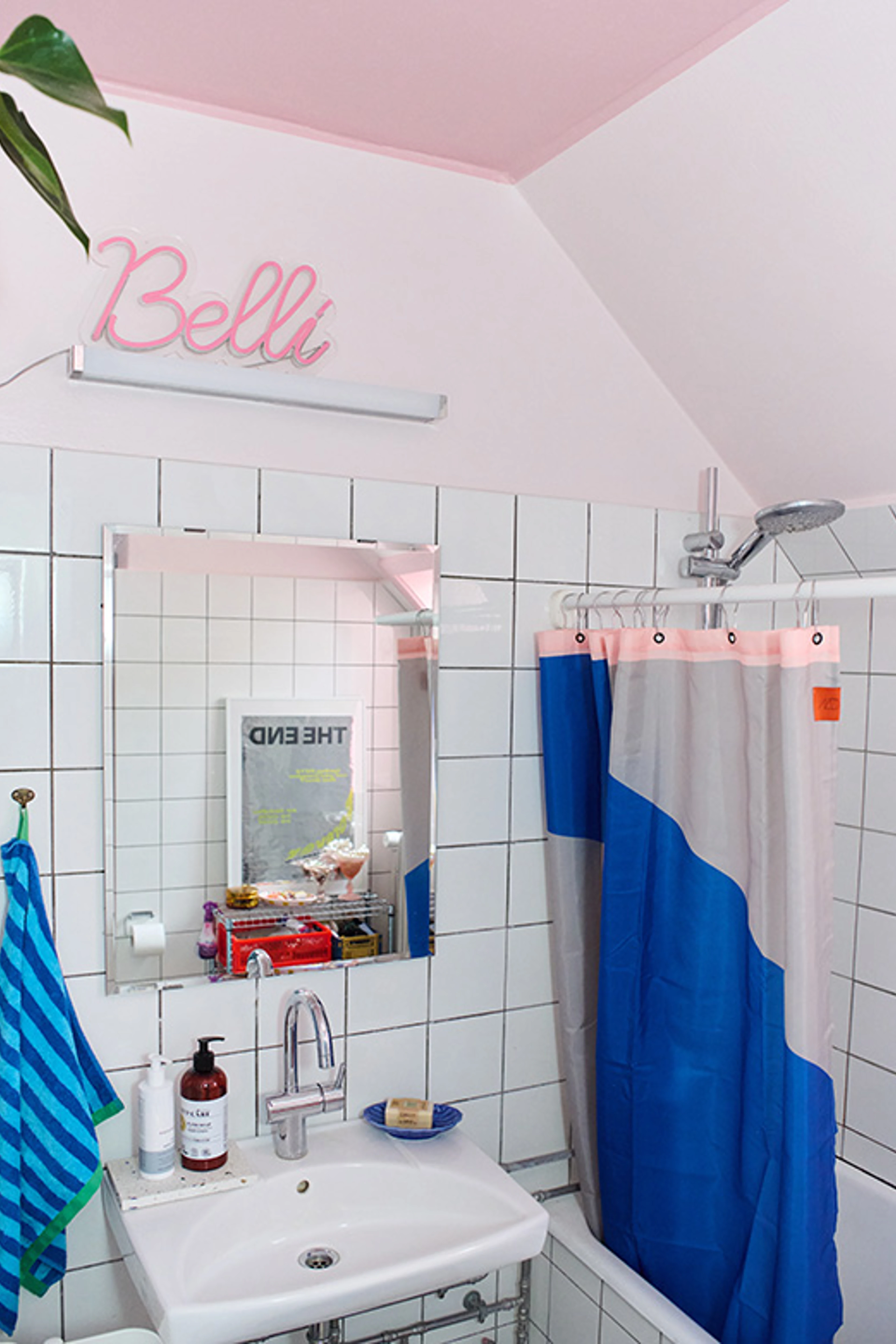 Badeværelse med hvide fliser og rosa loft, indrettet med vask, spejl, bruseforhæng og farverige detaljer.