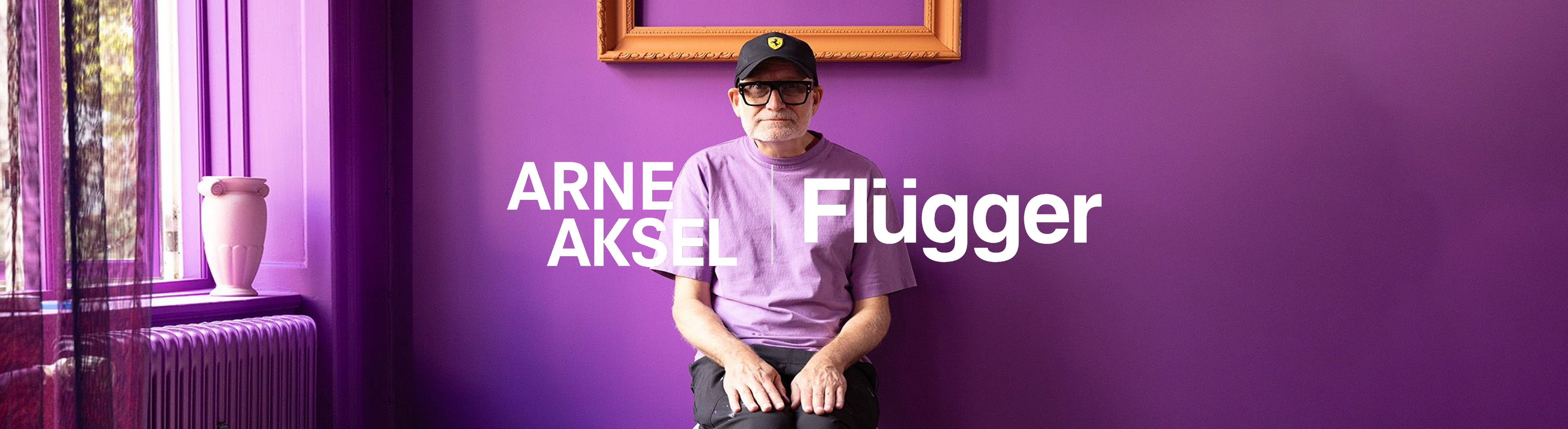 Stue med lilla vægge, hvor der sidder en hvidskægget mand. Manden har en lilla t-shirt, en sort kasket og sorte briller på.