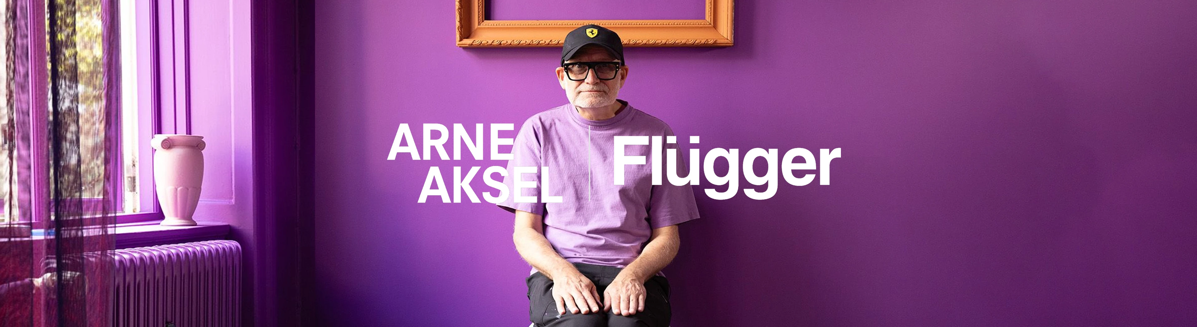 Stue med lilla vægge, hvor der sidder en hvidskægget mand. Manden har en lilla t-shirt, en sort kasket og sorte briller på.