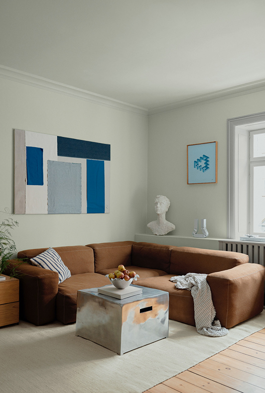 Stue med lyse vægge, brun sofa, kunst på væggene og sofabord med frugtskål.