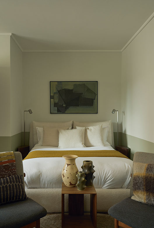 Calm bedroom with a double bed, light-colored textiles, a mustard yellow throw, abstract artwork, and ceramic vases.