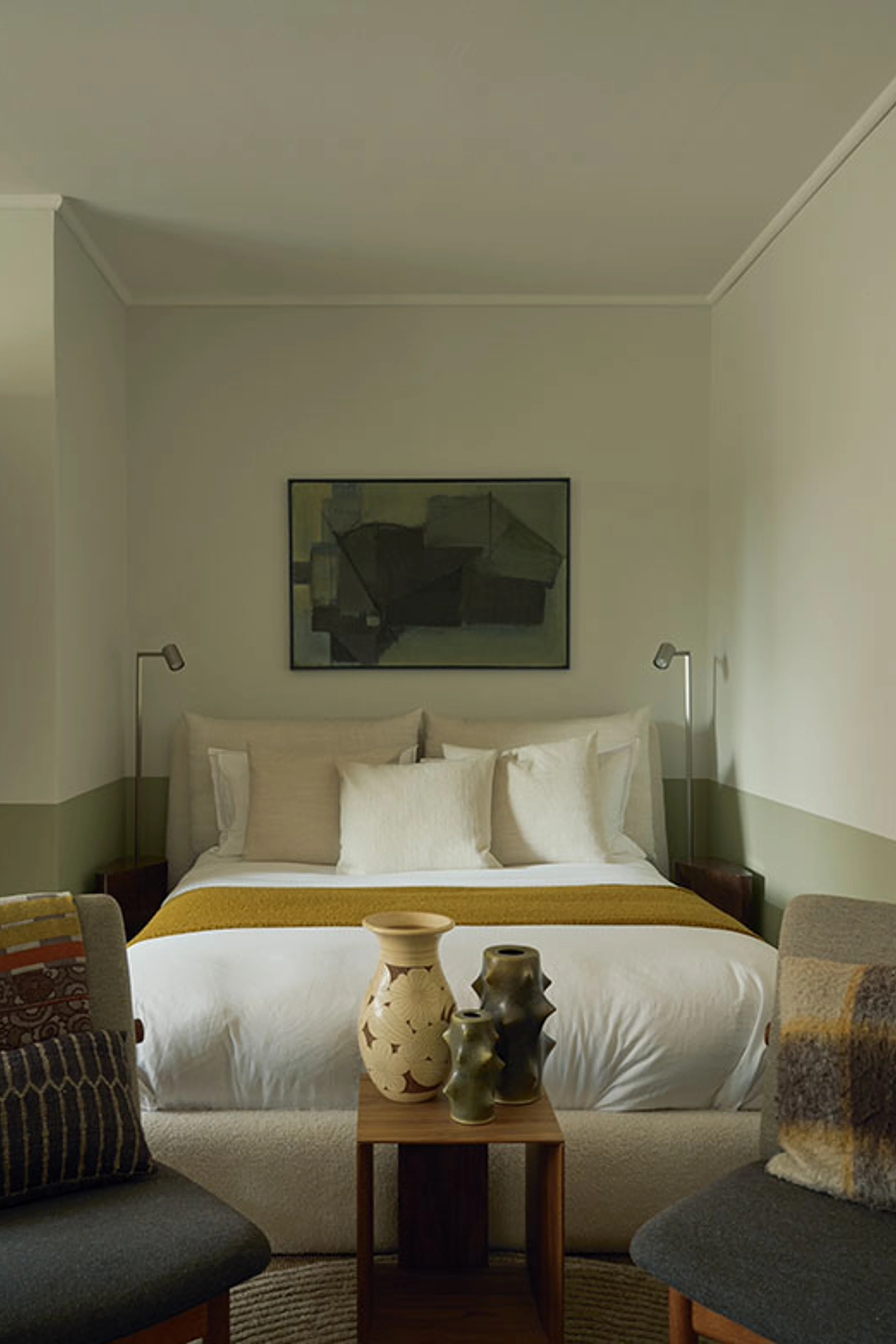 Calm bedroom with a double bed, light-colored textiles, a mustard yellow throw, abstract artwork, and ceramic vases.