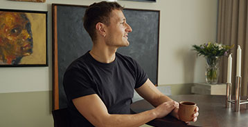 Man in a black T-shirt sits at a dark dining table, holding a mug and looking out the window.