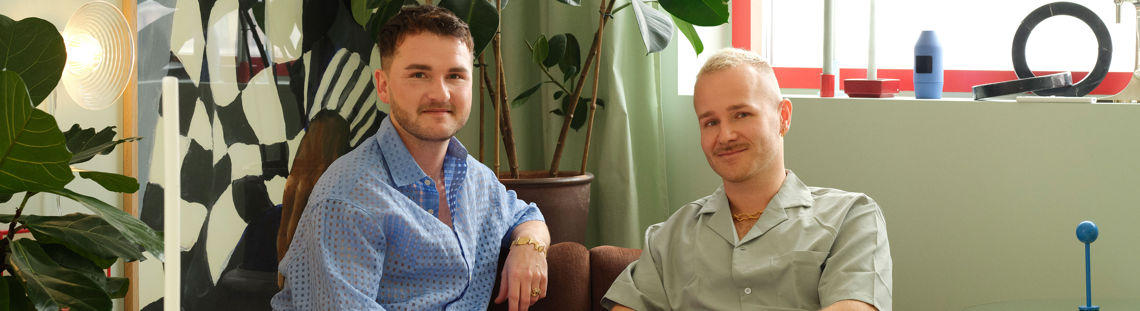 Two men sit together on a brown sofa in a colorful living room.