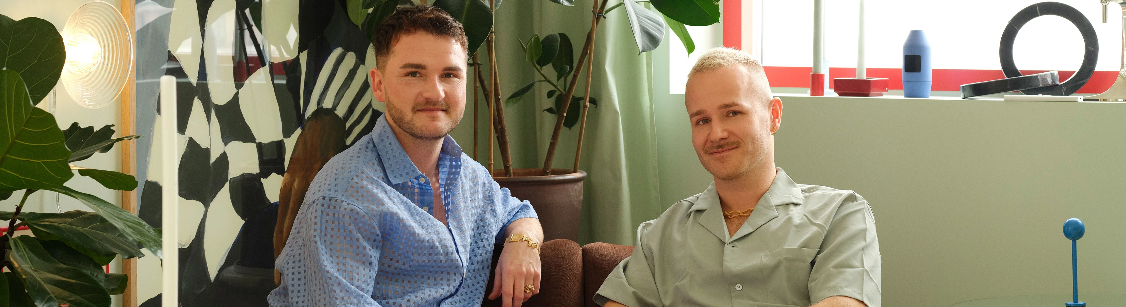 Two men sit together on a brown sofa in a colorful living room.