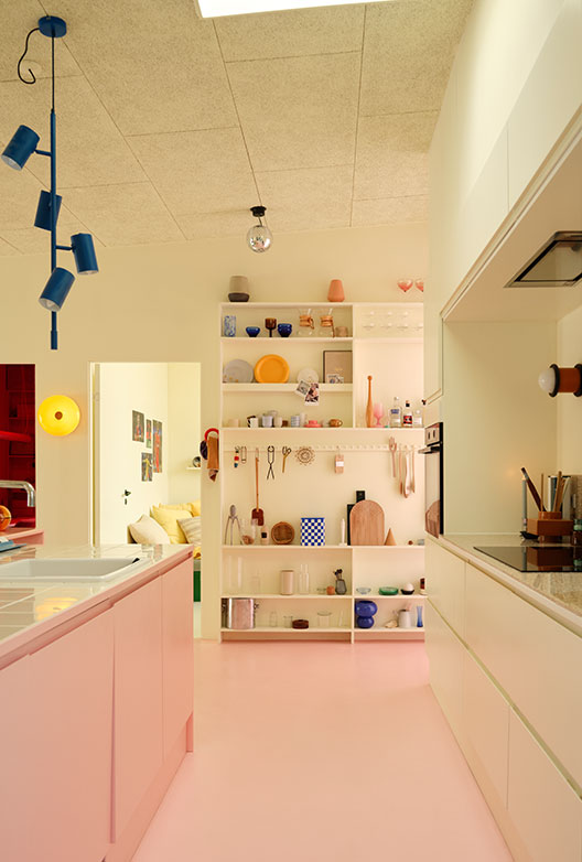 Colorful kitchen with a pink floor and kitchen island, light-colored cabinets, open shelving, and blue pendant lights.