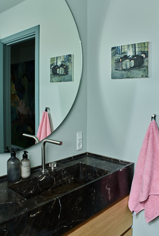 Blågrønt badeværelse med et rundt spejl, lyserødt håndklæde og en sort marmor vask. 