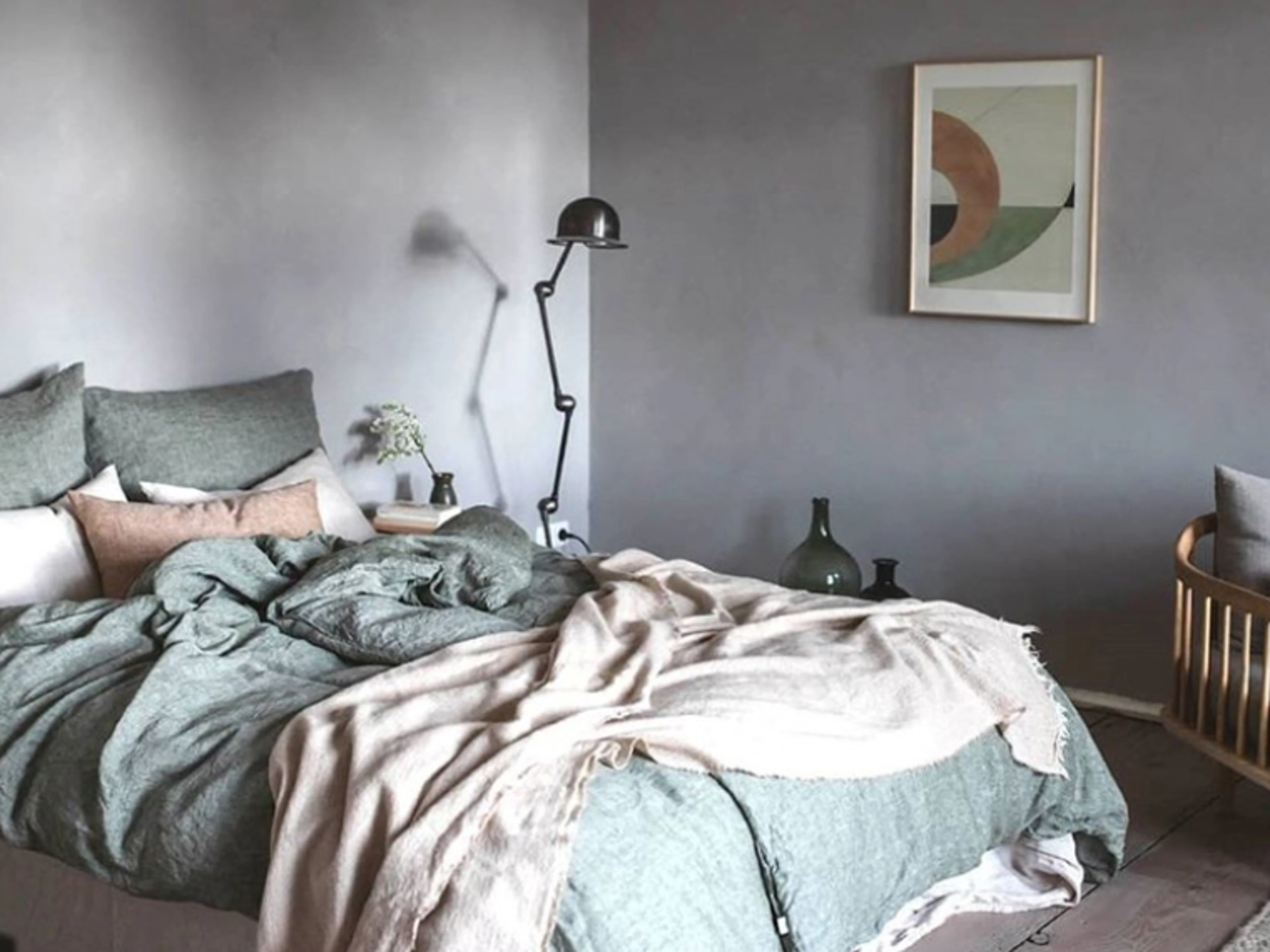 Soveværelse med vægge spartlet i grå, seng med grønne tekstiler og beige plaid, sort gulvlampe ved siden af sengen samt grafisk kunstværk på væggen.