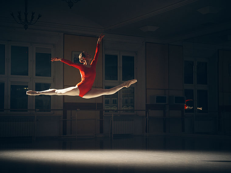 Balletdanser i rød dragt springer gennem luften i spagat i et mørkt dansestudie.