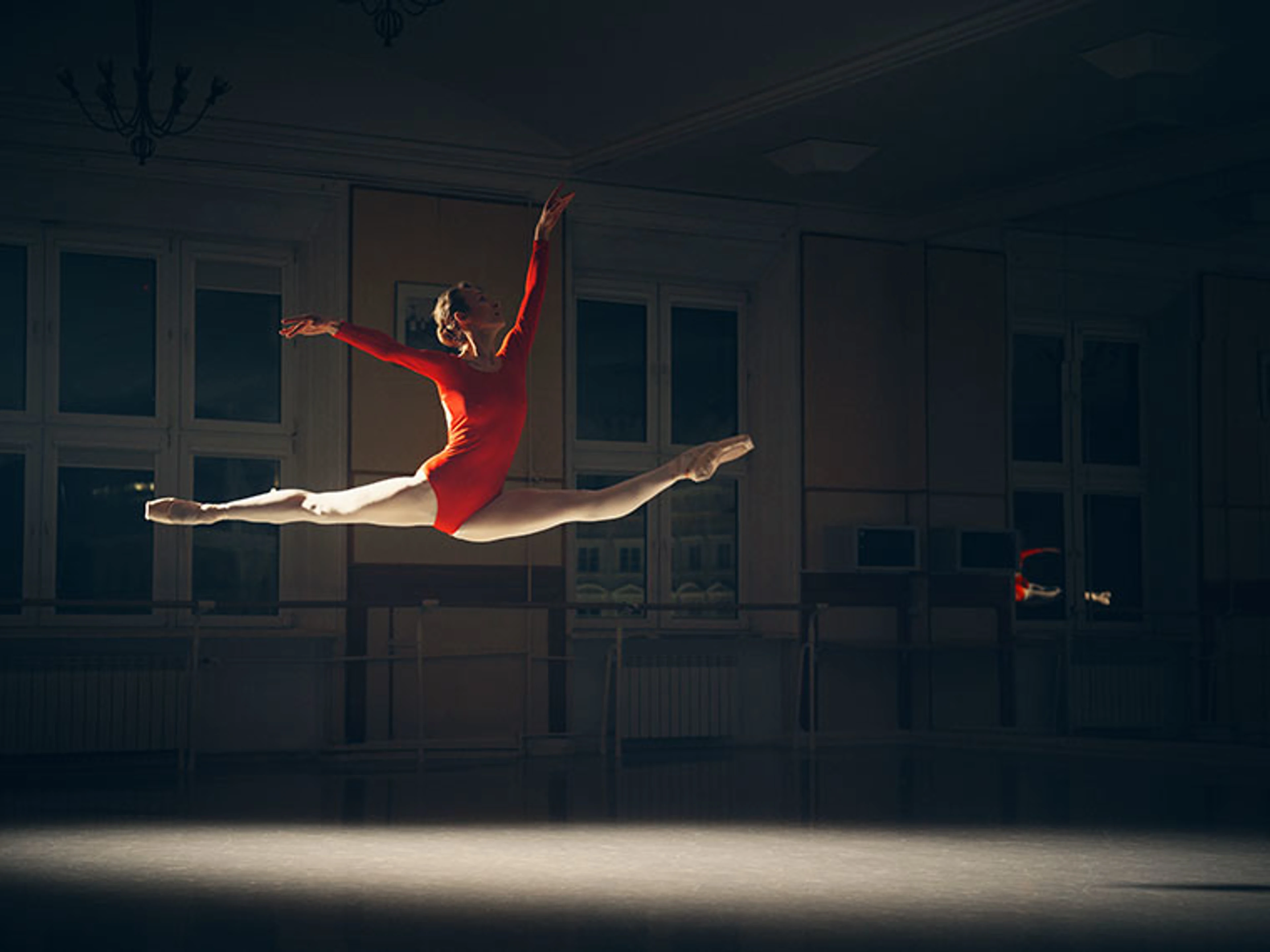 Balletdanser i rød dragt springer gennem luften i spagat i et mørkt dansestudie.