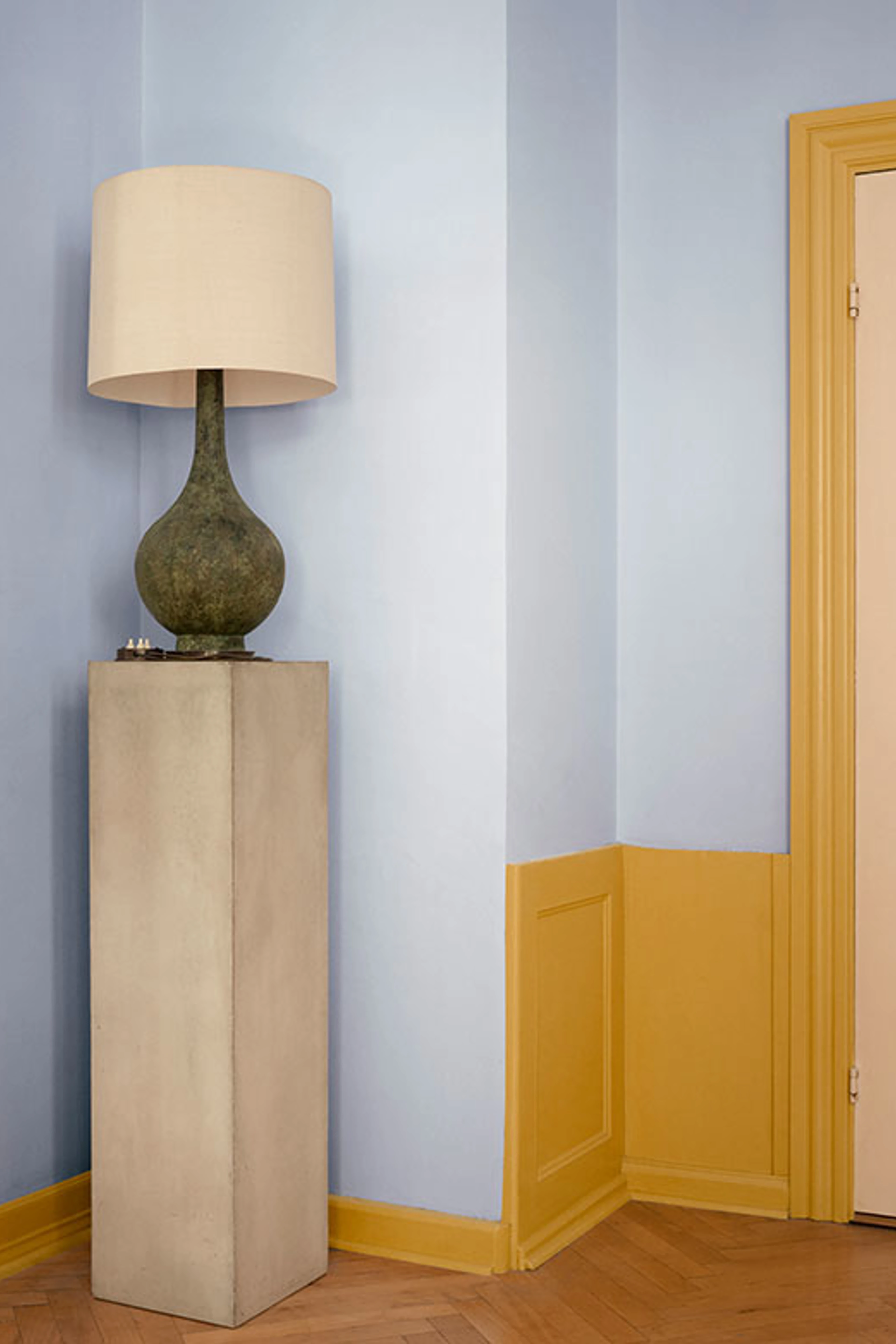 Interiør med lyseblå vægge og gul okkerfarvet panelbeklædning samt dør. En dekorativ bordlampe står på en høj sokkel i et klassisk rum med sildebensparket.