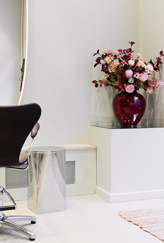 Rød vase med blomster på piedestal ved frisørplads i lys salon.